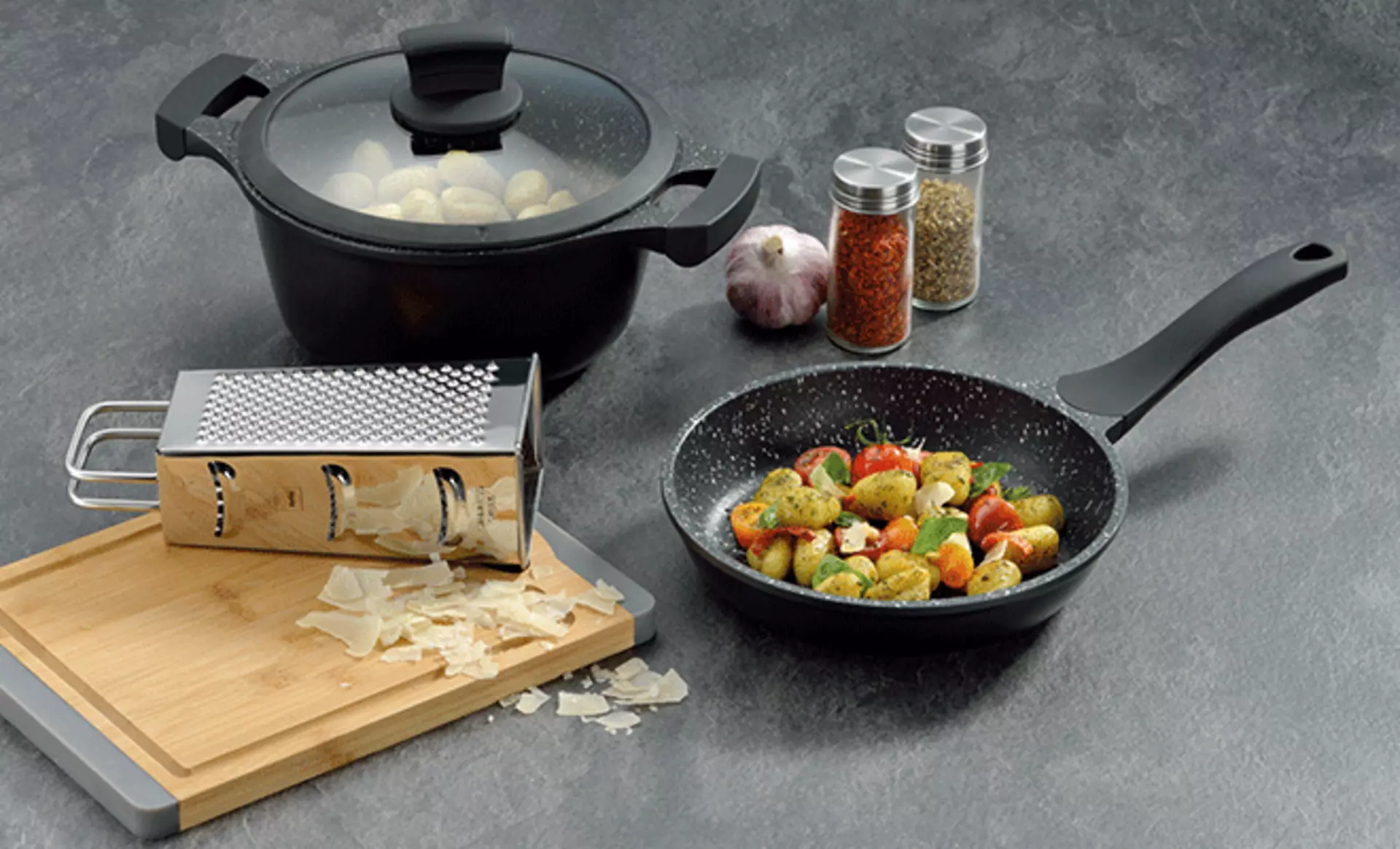 Eine Pfanne ein Topf und weitere Küchenhelfer auf grauem Hintergrund