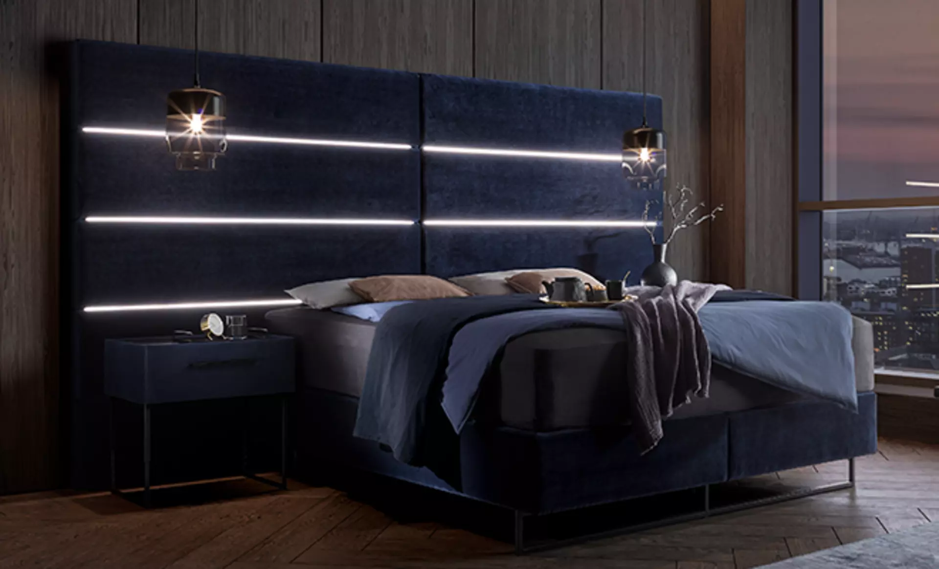 Ein sehr großes luxuriöses Boxspringbett mit dunkelblauen Bezügen und LED Streifen im sehr großen Kopfteil dekoriert mit Kissen und Plaids