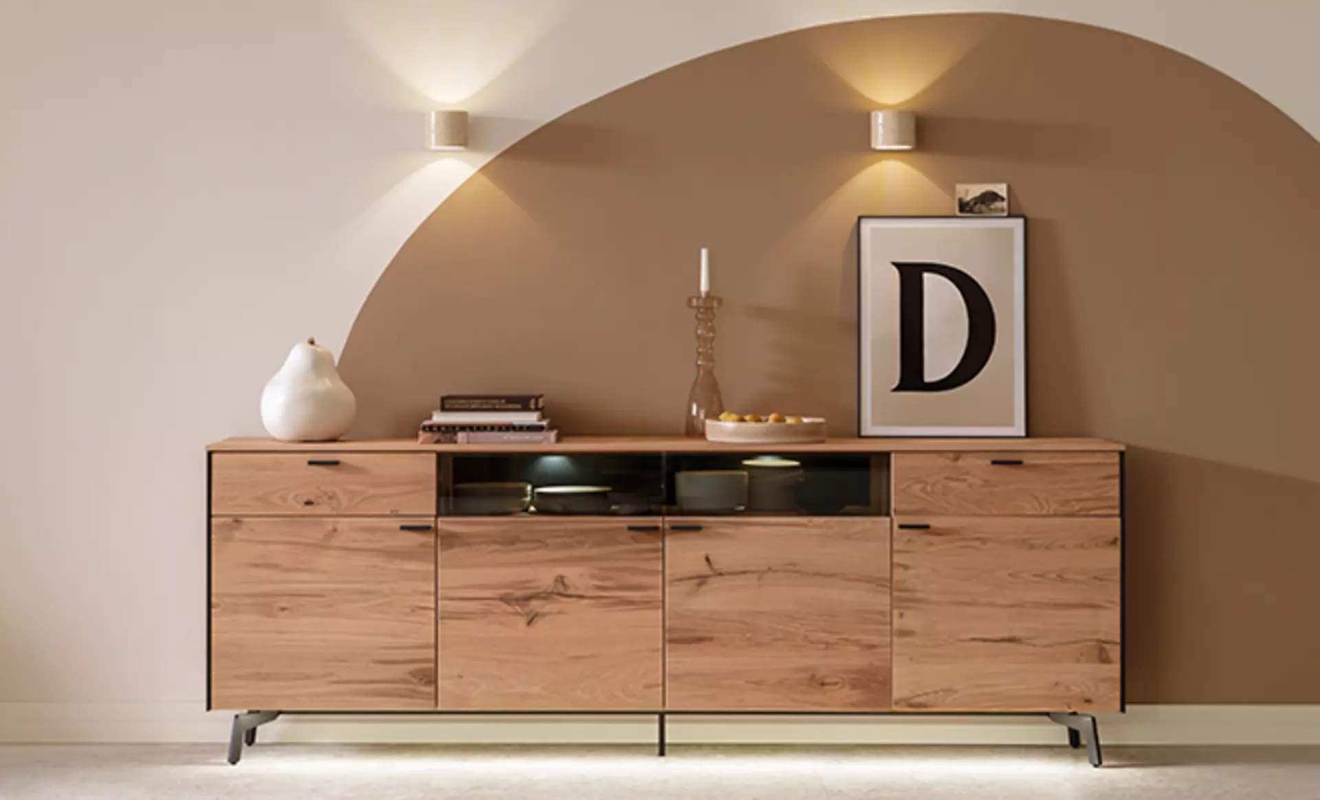 Das Andra Sideboard der Schöner Wohnen Kollektion aus massivem Holz mit Glas-Elementen an einer beleuchteten Wand mit beiger und brauner Wandfarbe