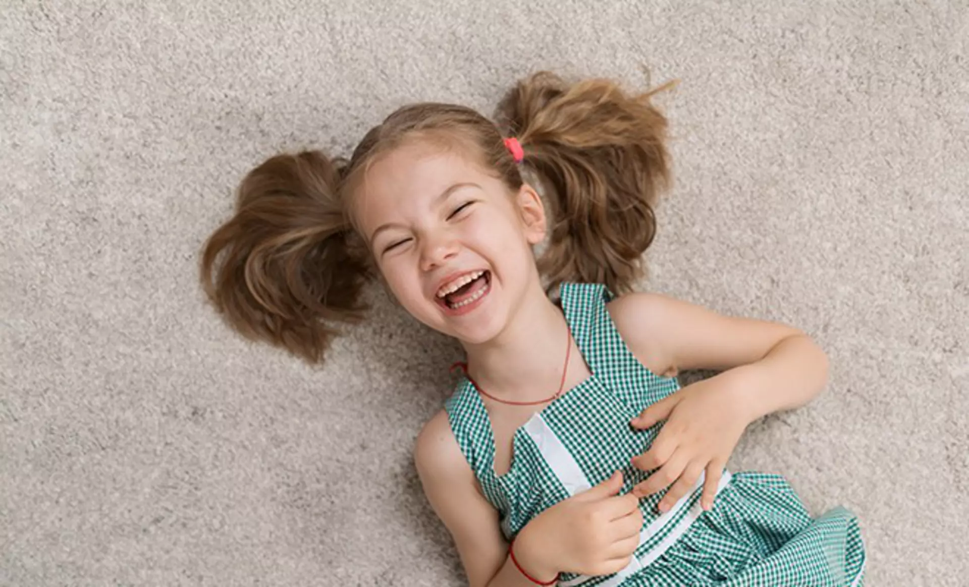 Kind sitzt lächelnd auf einem weichen grauen Teppich.