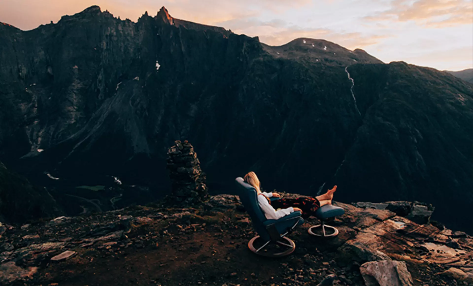 Landschaftsaufnahme der Norwegischen Berge. Eine Frau sitzt in einem Stressless Sessel an der Klippe und blickt in den Sonnenuntergang
