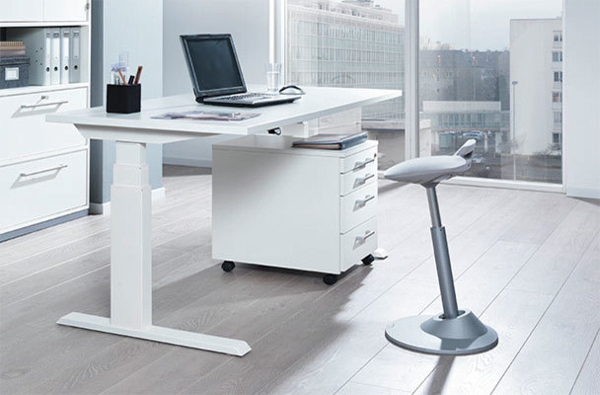 Weißer höhenverstellbarer Schreibtisch und davor ein grauer , höhenverstellbarer Sitzhocker in ergonomischer Form. Das Bild dient als zweites Milieubild für den Bereich Inspiration "Der richtige Bürostuhl".