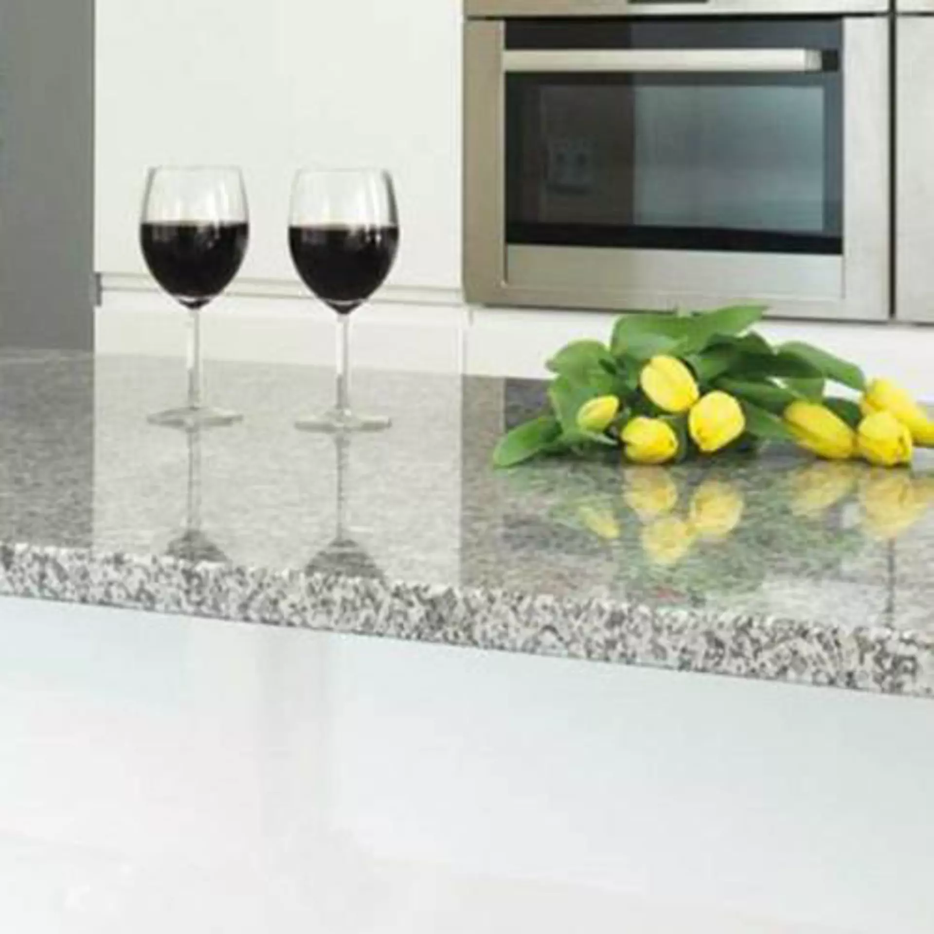Milieubild zu Arbeitsplatten aus Naturstein. Zu sehen ist eine Küchenzeile mit grau gesprenkeltem Naturstein. Darauf stehen zwei Gläser Rotwein und ein Bund gelber Tulpen.