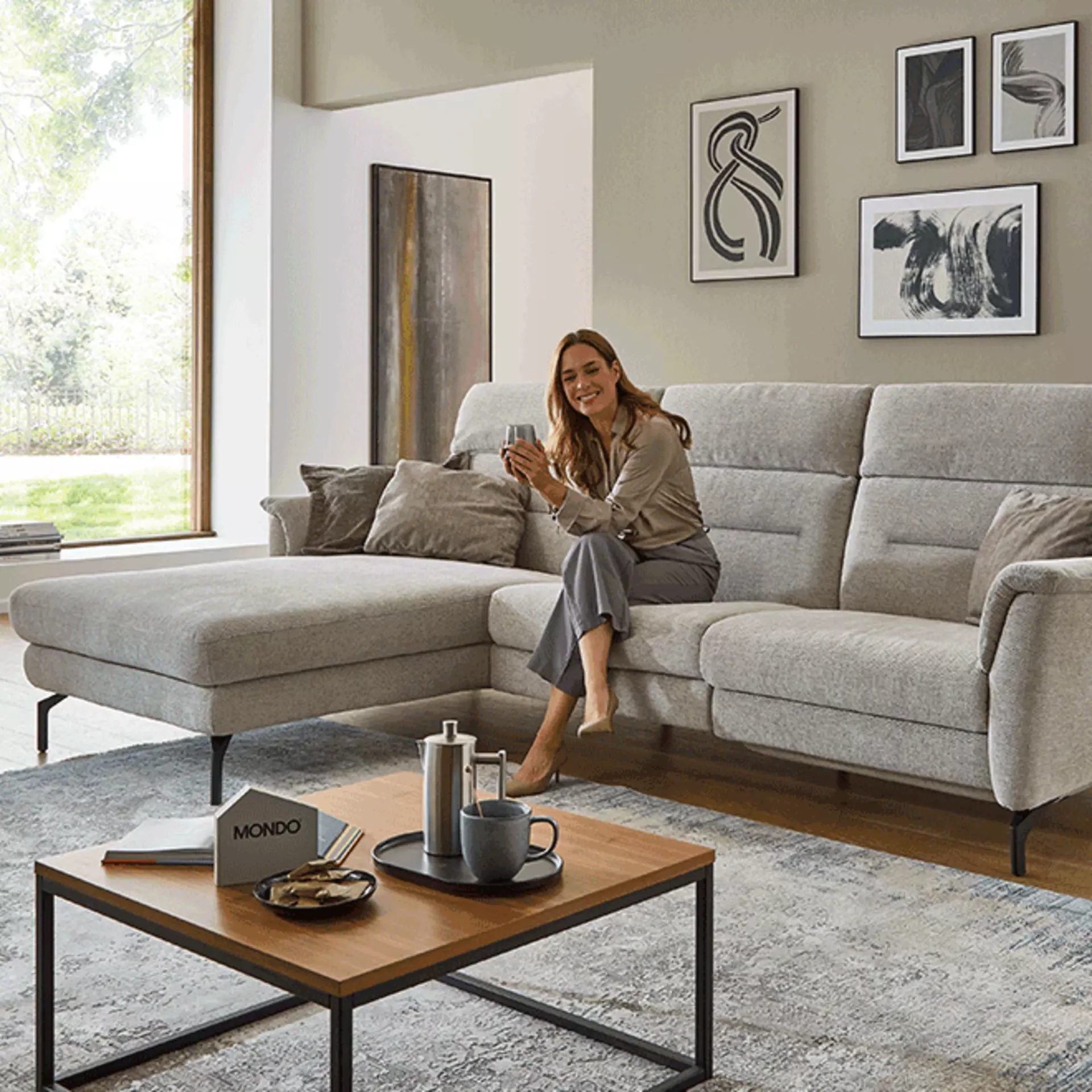 Frau im Wohnzimmer mit grauem Sofa und quadratischem Couchtisch aus Holz im schlichten Design