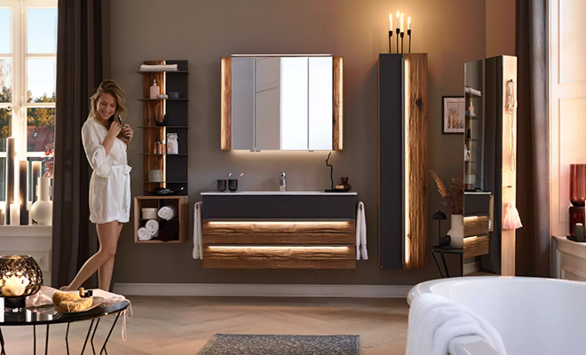 Die Jardin Badezimmer-Kollektion von Musterring mit rauem Massivholz und moderner LED Beleuchtung und schwarz matten akzenten. Eine Rau kämmt sich derweil die Haare