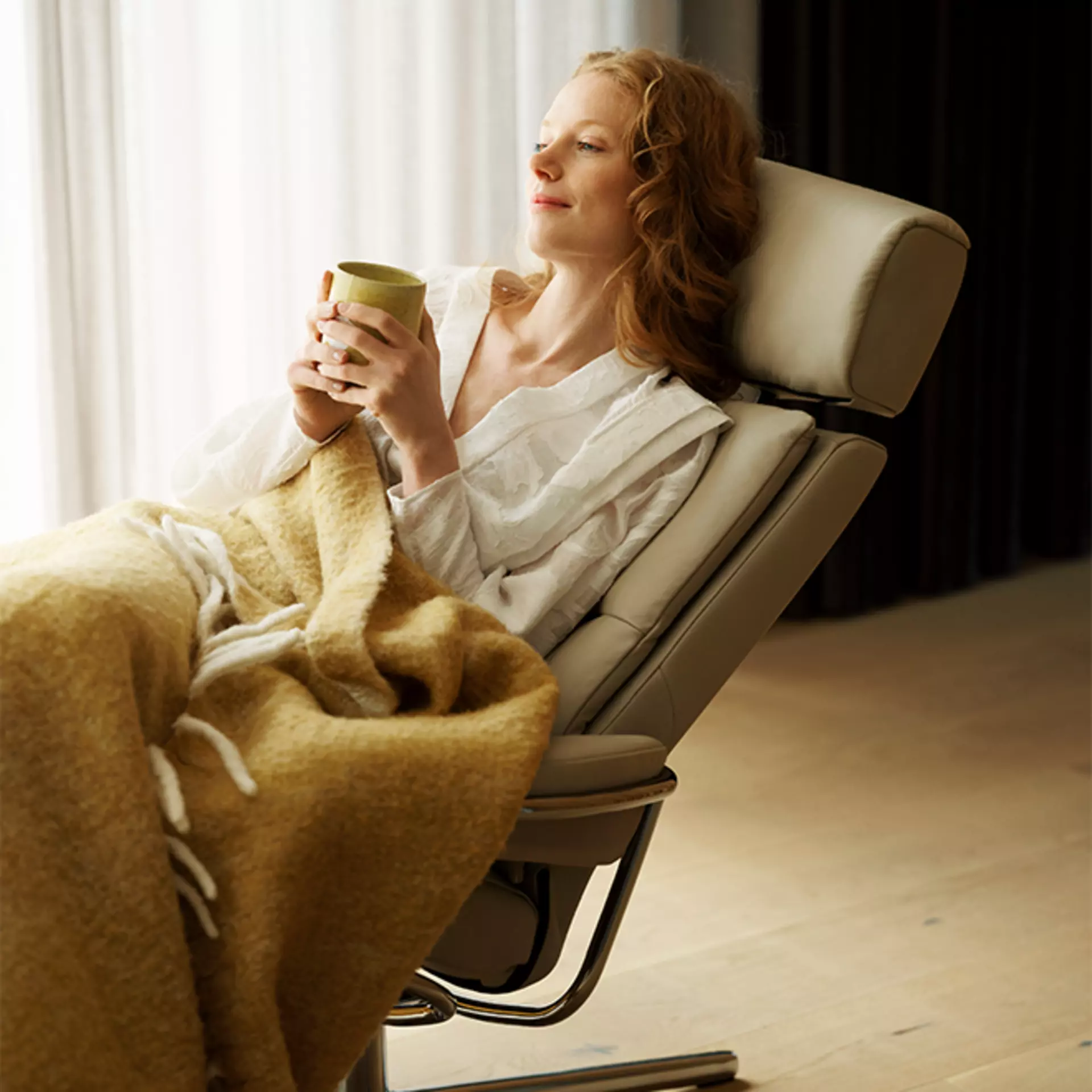eine Frau sitzt mit Kuscheldecke und Tasse Tee in der Hand in einem Stressless Tokyo Sessel mit Lederpolsterung