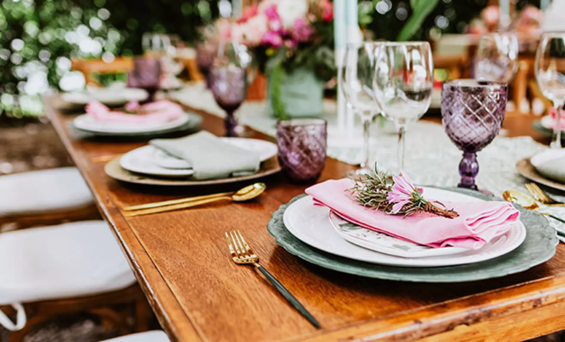 Esstisch aus Holz, dekoriert mit farbigen Tellern, klaren und violetten Gläsern, mit goldenem Besteck, rosa Servietten und frischen Blumen.