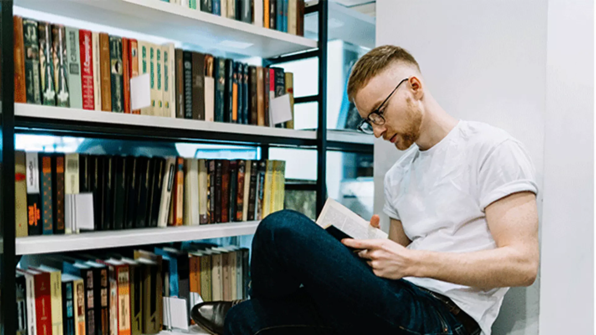 Junger Mann beim Lesen eines Buches neben einem Bücherregal