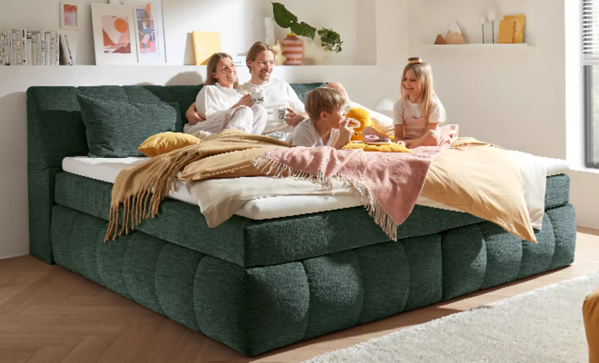 Sehr großes Familien-Boxspringbett in dunkelgrünem Stoffbezug dekoriert mit Tagesdecken. Die Familie mit zwei Kindern frühstückt darauf.