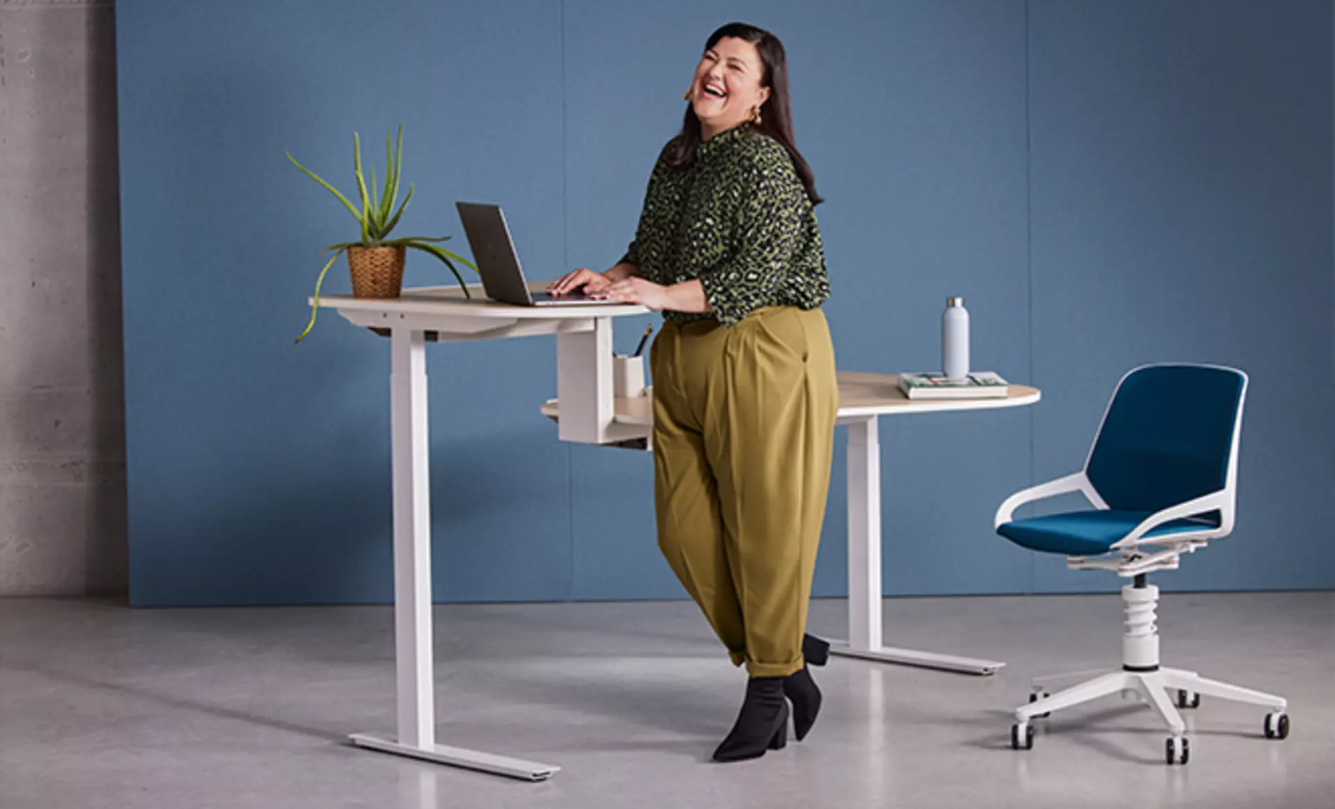Der höhenverstellbare aeris Active office Desk mit Holzfurnier. Davor steht der Numo bürostuhl mit blauem Stoffbezug und eine gutgelaunte Frau arbeitet am Steharbeitsplatz des Tisches