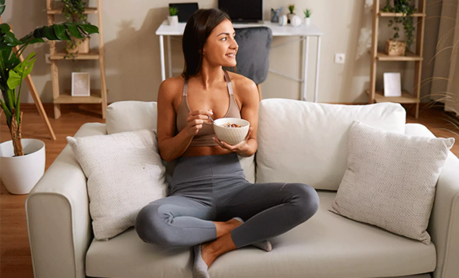 Eine Frau sitzt gemütlich auf ihrem Sofa und isst fröhlich ihr Frühstück.