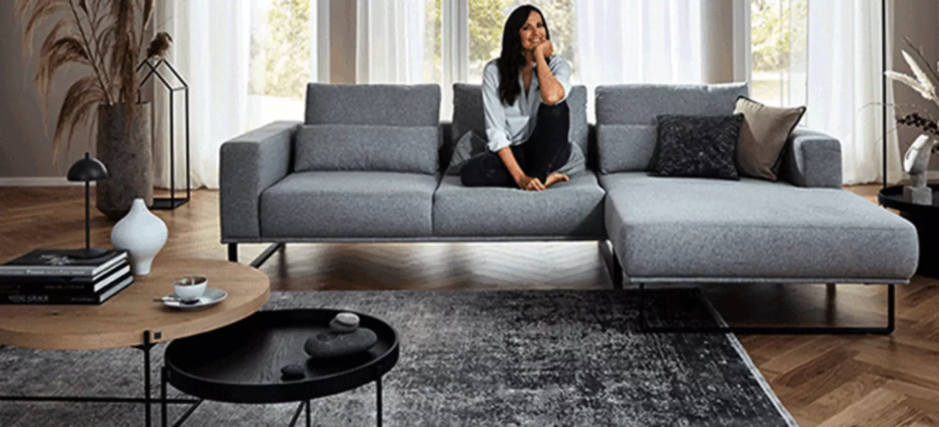 Die Schauspielerin Bettina Zimmermann auf einer grauen Couch in einem Wohnzimmer mit grauen und weißen Dekorationen.