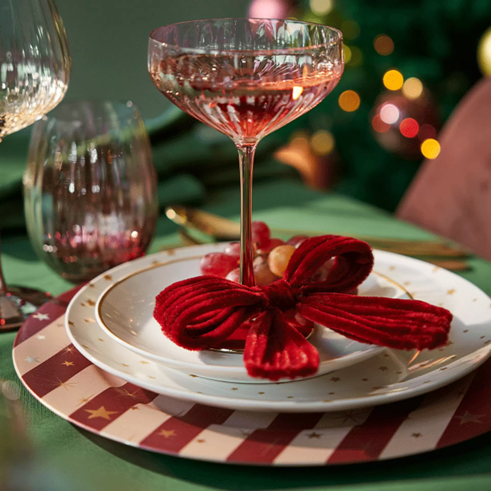 Weihnachtliche Platzdeko mit rot-weiß gestreiftem Set, Sektglas und rot gebundener Stoffschleife auf weißem Teller vor grünem Hintergrund