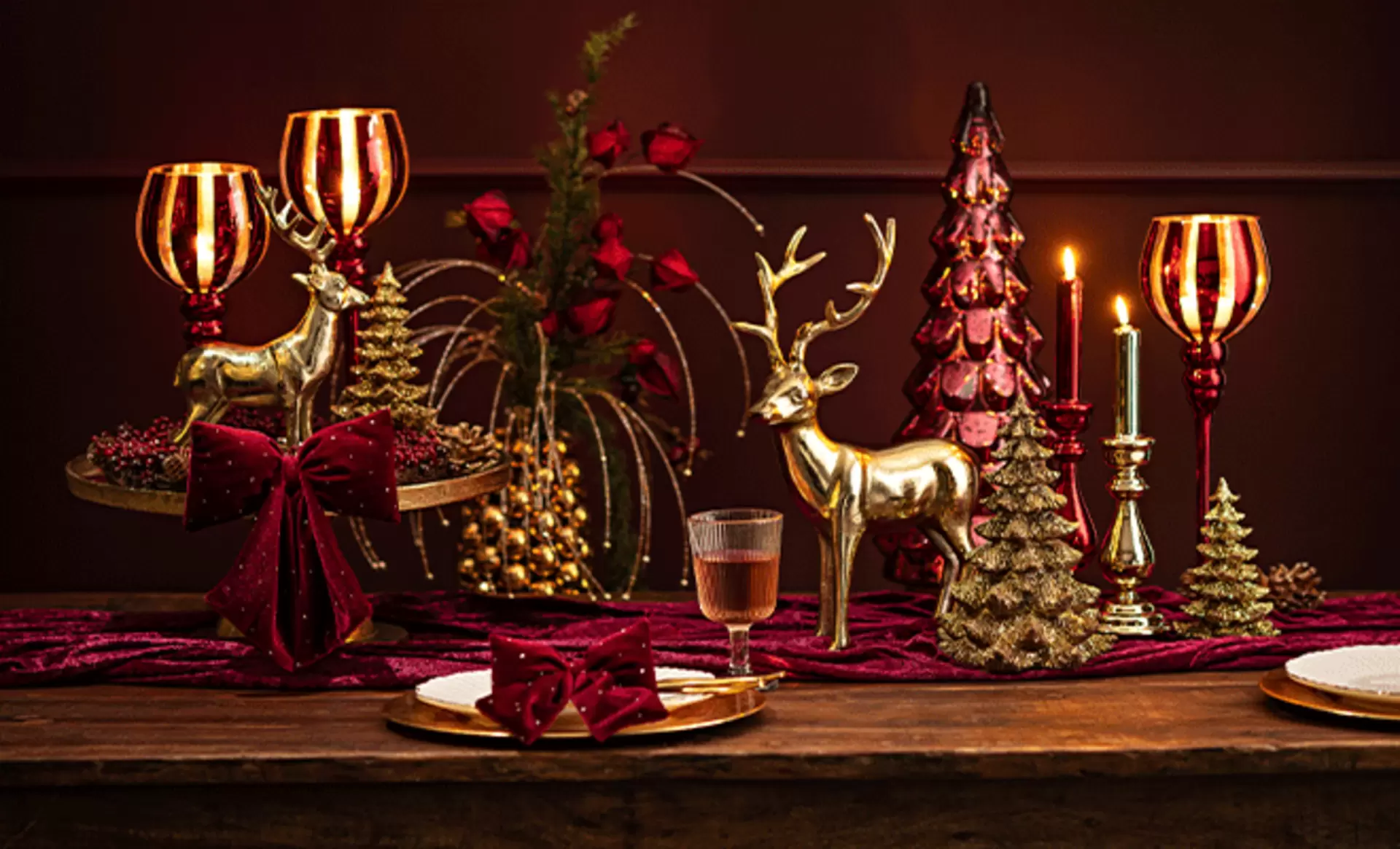 Elegant dekorierte weihnachtliche Festtafel mit goldenen Hirschfiguren, roten Windlichtern, Kerzenständern und kleiner Deko-Tanne auf dunkelrotem Läufer