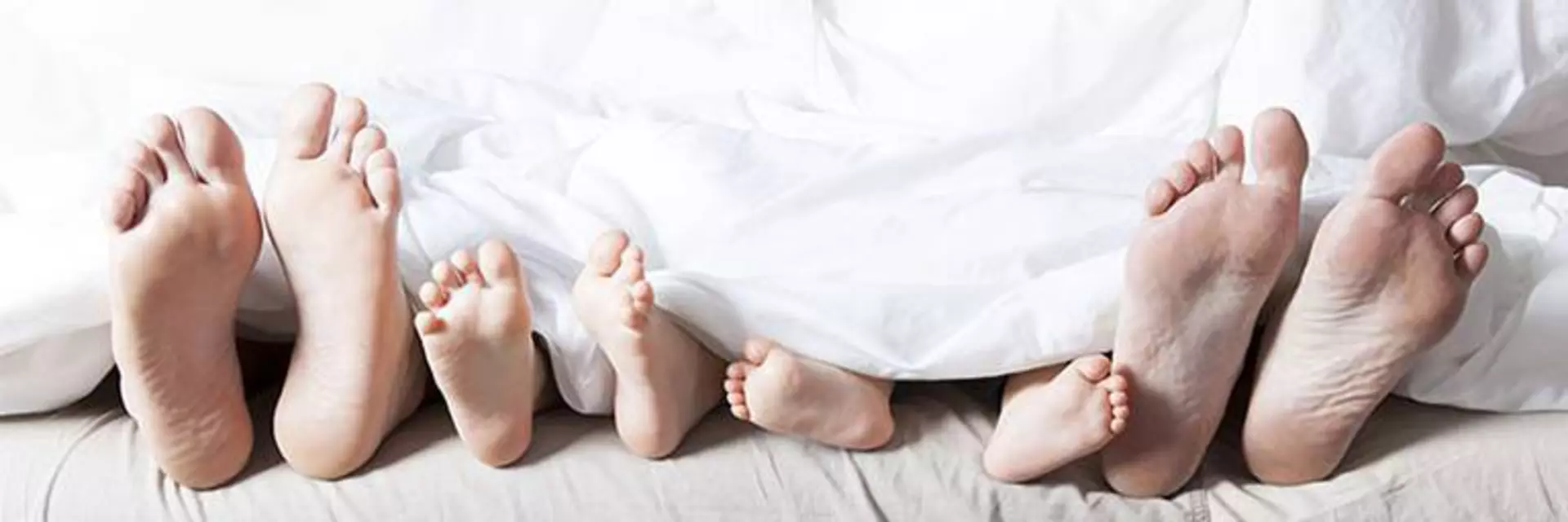 Vier Paar Füße schauen unter der weißen Bettdecke hervor. Es ist klar eine Familie, da es sich um Männerfüße, Frauenfüße und 2  Paar Kinderfüße handelt.