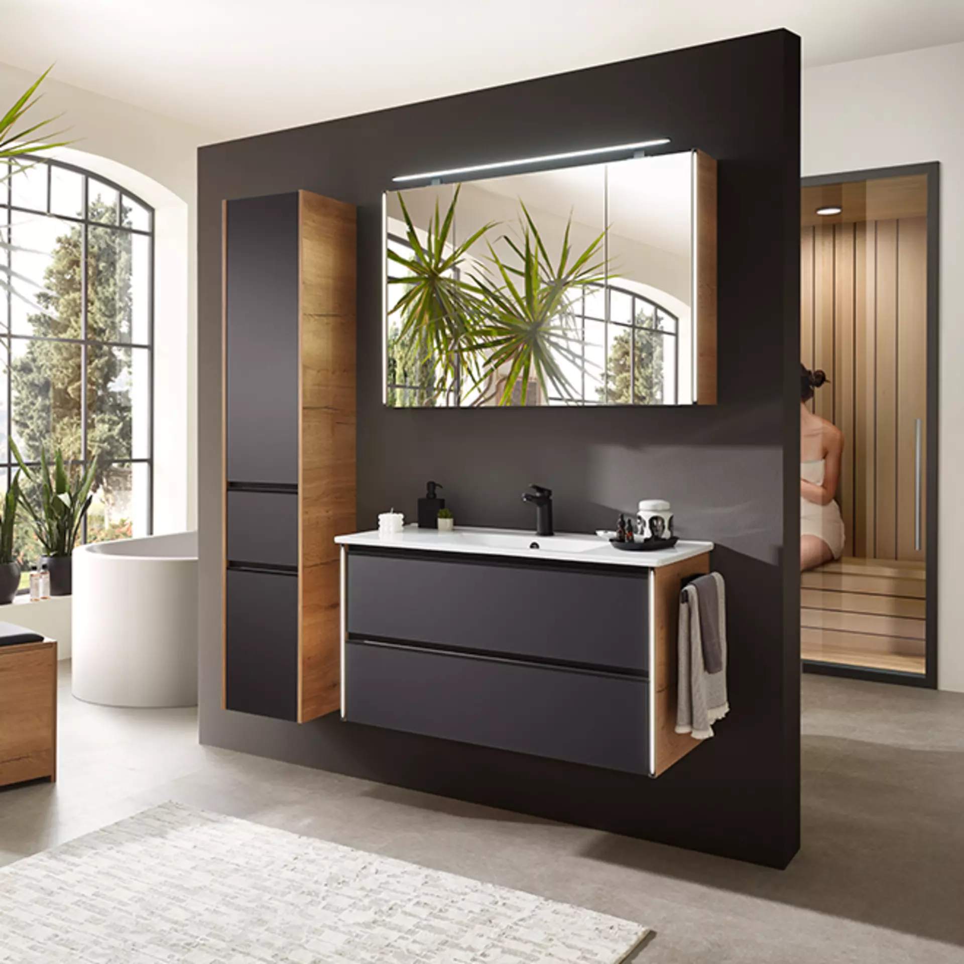 Die Serie 6040 Badezimmermöbel von Pelipal aus Eiche mit mattschwarzen Fronten mit Beleuchtung in einem großen offenen Badezimmer mit Sauna dahinter